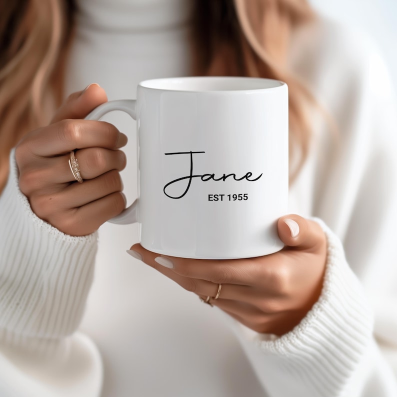 Puede incluir: Taza de cer&aacute;mica blanca con el nombre "Jane" en fuente cursiva y el texto "EST 1955" impreso en negro. La taza tiene un dise&ntilde;o simple y elegante, adecuada para caf&eacute; o t&eacute;.