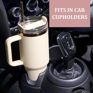 May include: Cream-colored travel mug with a gold rim and handle, placed in a car cupholder. The mug has a clear lid and a gold straw. The text "FITS IN CAR CUPHOLDERS" is displayed in a pink box above the mug.
