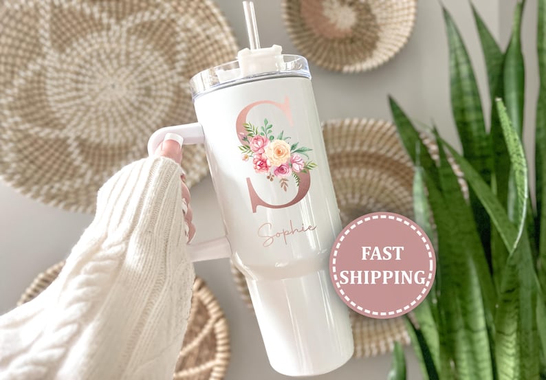 May include: White insulated tumbler with a handle, featuring a floral design and the letter "S" in rose gold. The name "Sophie" is written below the letter. Includes a straw and a white lid.