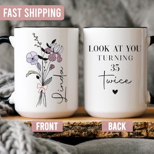 May include: White ceramic mug with a black handle and rim. One side features a floral bouquet with the name "Linda". The other side says "LOOK AT YOU TURNING 35 Twice" with a heart symbol. The mug is on a wooden surface.