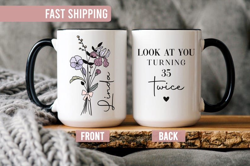 May include: White ceramic mug with a black handle and rim. One side features a floral bouquet with the name "Linda". The other side says "LOOK AT YOU TURNING 35 Twice" with a heart symbol. The mug is on a wooden surface.