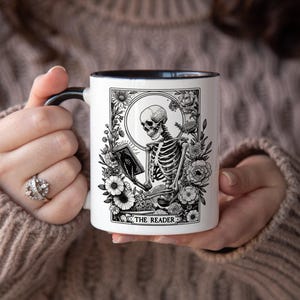 Puede incluir: Taza de cer&aacute;mica blanca con asa y borde negros, con una ilustraci&oacute;n en blanco y negro de un esqueleto leyendo un libro, rodeado de flores. El texto "THE READER" est&aacute; en la parte inferior. Un regalo &uacute;nico para los amantes de los libros.