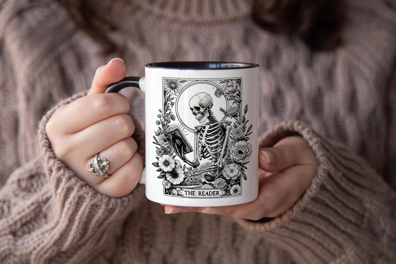 Puede incluir: Taza de cer&aacute;mica blanca con asa y borde negros, con una ilustraci&oacute;n en blanco y negro de un esqueleto leyendo un libro, rodeado de flores. El texto "THE READER" est&aacute; en la parte inferior. Un regalo &uacute;nico para los amantes de los libros.