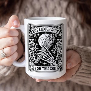 May include: White ceramic mug with a black and white graphic design. The design features a skeleton hand holding a bundle of sage, a crescent moon, roses, and the text "NOT ENOUGH SAGE FOR THIS SHIT."