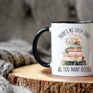 May include: White ceramic mug with a black handle and rim, featuring the text "THERE'S NO SUCH THING AS TOO MANY BOOKS." A stack of books with floral accents is illustrated on the front. The mug sits on a wooden surface.