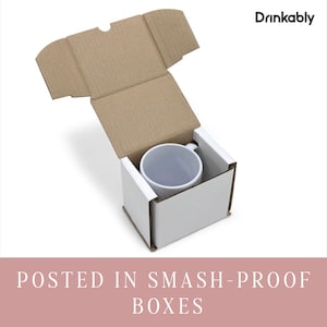 May include: A white ceramic mug inside a white box, nestled within a brown cardboard shipping box. The top of the shipping box is open. The text "Drinkably" is in the upper right corner. The text "POSTED IN SMASH-PROOF BOXES" is at the bottom.