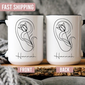 May include: Two white ceramic mugs with black handles and rims. Each mug features a black line drawing of a flower inside an ear shape, with the name "Hannah" below. The front and back of the mugs are labeled. The top banner says "FAST SHIPPING".