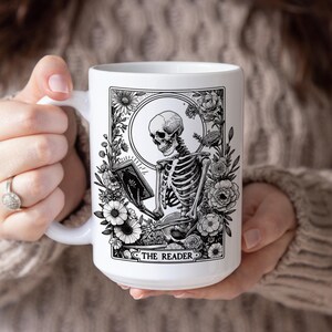 Puede incluir: Taza de cer&aacute;mica blanca con una ilustraci&oacute;n en blanco y negro de un esqueleto leyendo un libro, rodeado de flores y follaje. El texto "THE READER" est&aacute; en la parte inferior. La taza es sostenida por una persona.