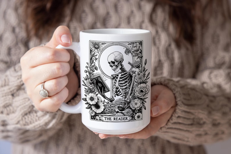 Puede incluir: Taza de cer&aacute;mica blanca con una ilustraci&oacute;n en blanco y negro de un esqueleto leyendo un libro, rodeado de flores y follaje. El texto "THE READER" est&aacute; en la parte inferior. La taza es sostenida por una persona.