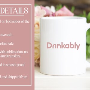 May include: White ceramic mug with the word "Drinkably" in a soft pink font. The mug has a curved handle and is set against a backdrop of flowers and greenery. The mug is microwave and dishwasher safe.