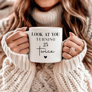 May include: A white ceramic mug featuring the black printed text LOOK AT YOU TURNING 25 twice with a small black heart symbol centered below the text. The mug has a smooth finish and a standard handle, held by two hands with manicured fingernails.