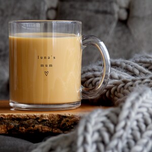 May include: A clear glass mug filled with a light brown liquid, possibly coffee or tea. The mug has a handle and the words "luna's mum" with a heart symbol are printed on the side. The mug sits on a wooden surface.