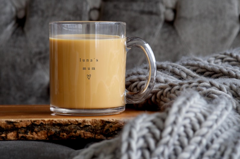 May include: A clear glass mug filled with a light brown liquid, possibly coffee or tea. The mug has a handle and the words "luna's mum" with a heart symbol are printed on the side. The mug sits on a wooden surface.