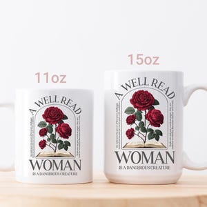 May include: Two white ceramic mugs, 11oz and 15oz, with a floral design. The design features red roses, an open book, and the text "A WELL READ WOMAN IS A DANGEROUS CREATURE". The mugs have a curved handle and are set on a wooden surface.