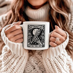 May include: White ceramic mug with a black and white graphic design. The design features a skeleton hand holding a bundle of sage, a crescent moon, and the text "NOT ENOUGH SAGE FOR THIS SHIT". The mug is held by a person wearing a cream-colored sweater.