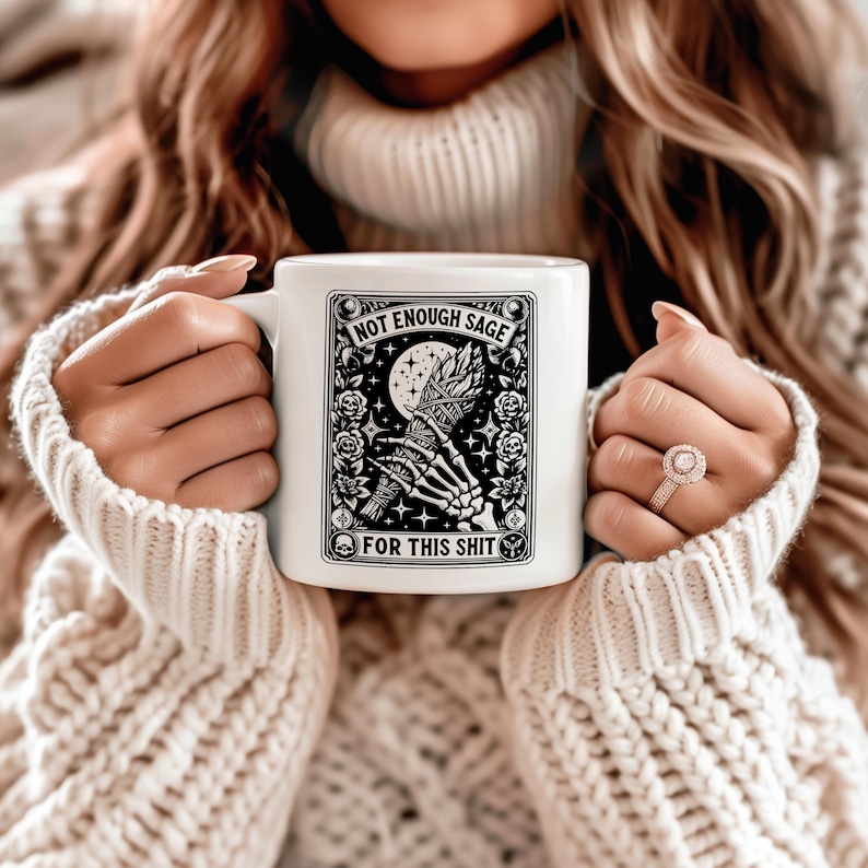 May include: White ceramic mug with a black and white graphic design. The design features a skeleton hand holding a bundle of sage, a crescent moon, and the text "NOT ENOUGH SAGE FOR THIS SHIT". The mug is held by a person wearing a cream-colored sweater.