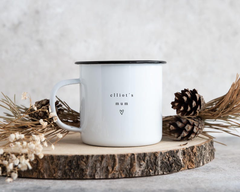 May include: White enamel mug with a black rim and handle. The mug has the text "elliot's mum" and a heart symbol printed on the front. The mug is displayed on a wooden slice with pine cones and dried foliage.