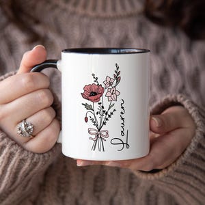 May include: White ceramic mug with a black handle and rim, featuring a floral bouquet design in pink and green. The word "Lauren" is written vertically in a script font. The mug is held by a person wearing a brown sweater.