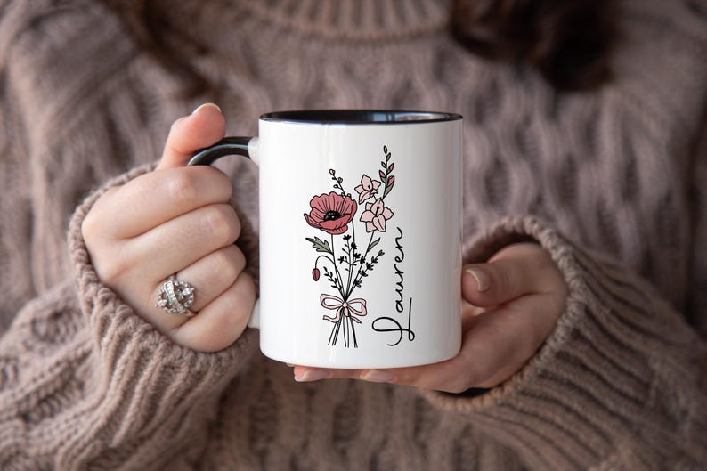 May include: White ceramic mug with a black handle and rim, featuring a floral bouquet design in pink and green. The word "Lauren" is written vertically in a script font. The mug is held by a person wearing a brown sweater.