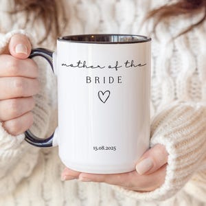 May include: White ceramic mug with a black interior and handle. The mug features the text "mother of the BRIDE" in a script font, a heart symbol, and the date "15.08.2025". The mug is held by a person wearing a white sweater.