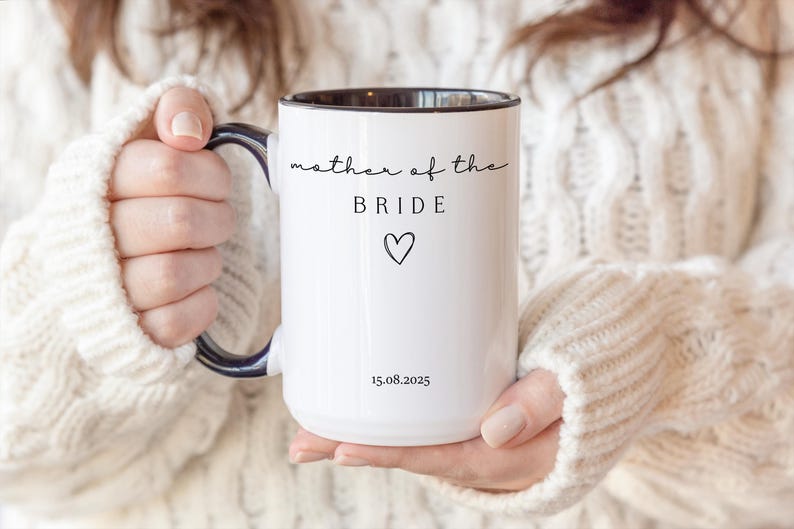 May include: White ceramic mug with a black interior and handle. The mug features the text "mother of the BRIDE" in a script font, a heart symbol, and the date "15.08.2025". The mug is held by a person wearing a white sweater.