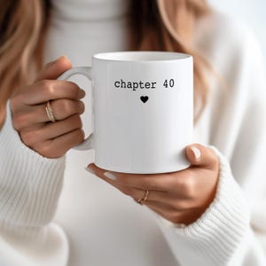 May include: White ceramic mug with the text "chapter 40" and a black heart symbol. The mug is held by a person wearing a white sweater. The mug is a standard size and has a curved handle.
