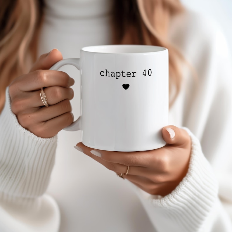 May include: White ceramic mug with the text "chapter 40" and a black heart symbol. The mug is held by a person wearing a white sweater. The mug is a standard size and has a curved handle.