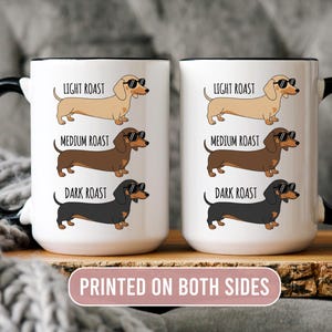 May include: Two white coffee mugs with black handles and rims, each featuring a dachshund illustration in light, medium, and dark roast colors, wearing sunglasses. The mugs have the text "LIGHT ROAST", "MEDIUM ROAST", and "DARK ROAST". The text "PRINTED ON BOTH SIDES" is also visible.