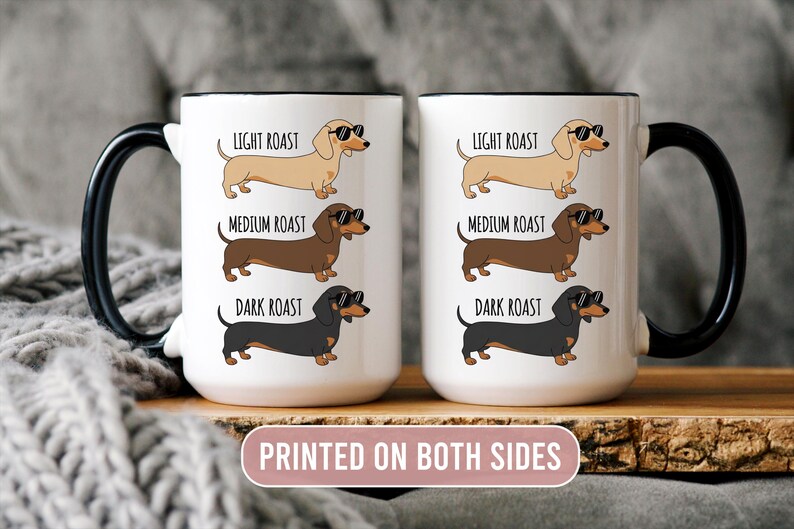 May include: Two white coffee mugs with black handles and rims, each featuring a dachshund illustration in light, medium, and dark roast colors, wearing sunglasses. The mugs have the text "LIGHT ROAST", "MEDIUM ROAST", and "DARK ROAST". The text "PRINTED ON BOTH SIDES" is also visible.
