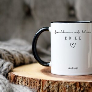 May include: White ceramic mug with a black handle and rim. The mug features the text "father of the BRIDE" in a delicate script, along with a heart symbol and the date 15.08.2025. The mug is set on a wooden surface.
