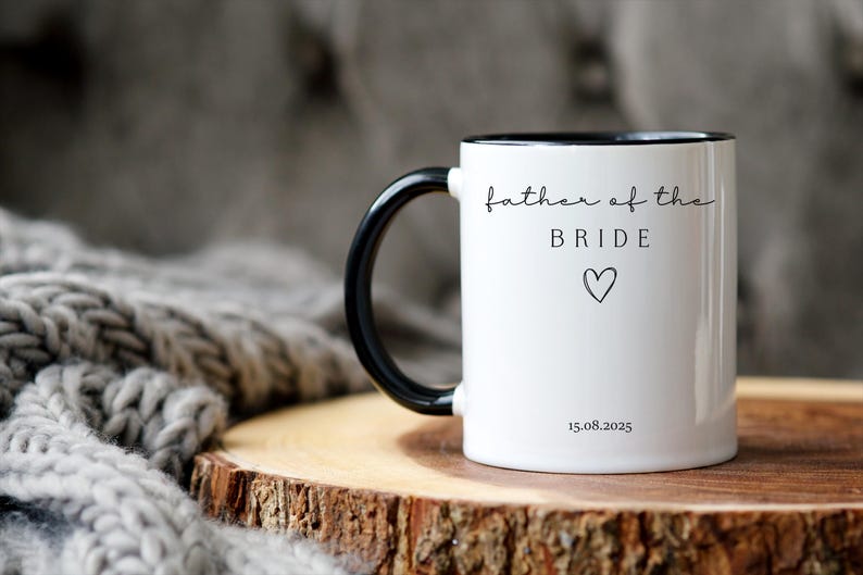 May include: White ceramic mug with a black handle and rim. The mug features the text "father of the BRIDE" in a delicate script, along with a heart symbol and the date 15.08.2025. The mug is set on a wooden surface.