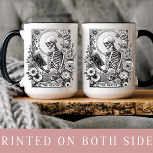 Puede incluir: Dos tazas de cer&aacute;mica blanca con asas y bordes negros. Cada taza presenta una ilustraci&oacute;n en blanco y negro de un esqueleto leyendo un libro, rodeado de flores, con el texto "THE READER" en la parte inferior. Las tazas est&aacute;n sobre una superficie de madera.