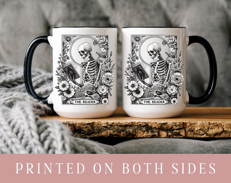 Puede incluir: Dos tazas de cer&aacute;mica blanca con asas y bordes negros. Cada taza presenta una ilustraci&oacute;n en blanco y negro de un esqueleto leyendo un libro, rodeado de flores, con el texto "THE READER" en la parte inferior. Las tazas est&aacute;n sobre una superficie de madera.