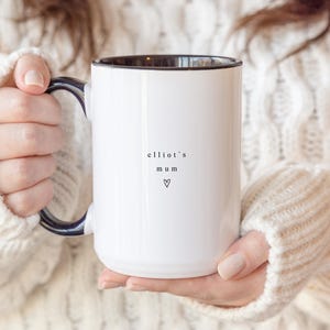 May include: White ceramic mug with a black interior and handle. The mug features the text "elliot's mum" and a heart symbol. The mug is held by a person wearing a white cable-knit sweater.