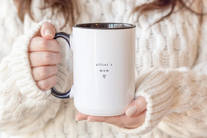 May include: White ceramic mug with a black interior and handle. The mug features the text "elliot's mum" and a heart symbol. The mug is held by a person wearing a white cable-knit sweater.