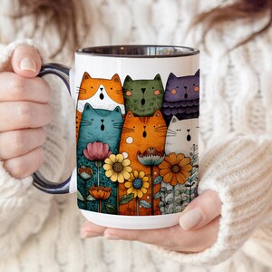 Może przedstawiać: Biały ceramiczny kubek z kolorową ilustracją stylizowanych kot&oacute;w i kwiat&oacute;w. Koty są w r&oacute;żnych kolorach, w tym pomarańczowym, niebieskim, zielonym i fioletowym. Kwiaty są ż&oacute;łte, pomarańczowe i r&oacute;żowe. Kubek ma czarny uchwyt i rant.