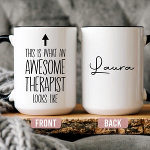 May include: A white ceramic mug with a black handle and the text "But First, Coffee" in a black, handwritten-style font. The mug is on a white surface.