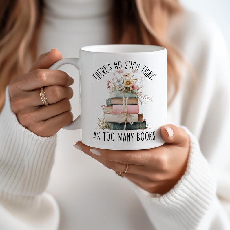 May include: White ceramic mug with the text "THERE'S NO SUCH THING AS TOO MANY BOOKS" and a stack of books with flowers. The mug has a handle and is held by a person. The books are in shades of blue, pink, and green.