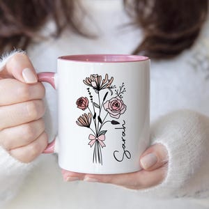 May include: A white ceramic mug with a pink interior and handle, featuring a black line art illustration of a floral bouquet tied with a pink bow and the name Sarah written in black cursive script on the side of the mug.