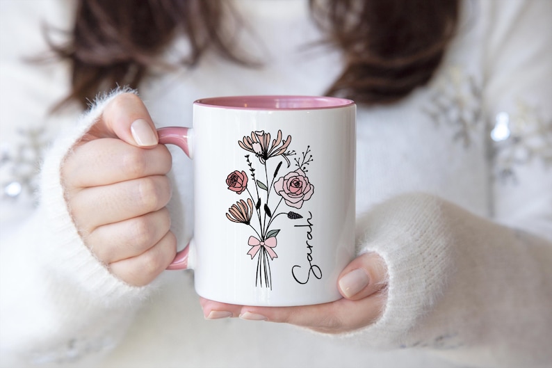 May include: A white ceramic mug with a pink interior and handle, featuring a black line art illustration of a floral bouquet tied with a pink bow and the name Sarah written in black cursive script on the side of the mug.