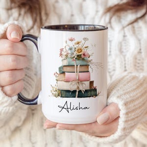 May include: White ceramic mug with a black rim and handle, featuring a stack of books tied with twine and topped with flowers. The name "Alisha" is written in script on the front. The mug is held by a person wearing a white sweater.