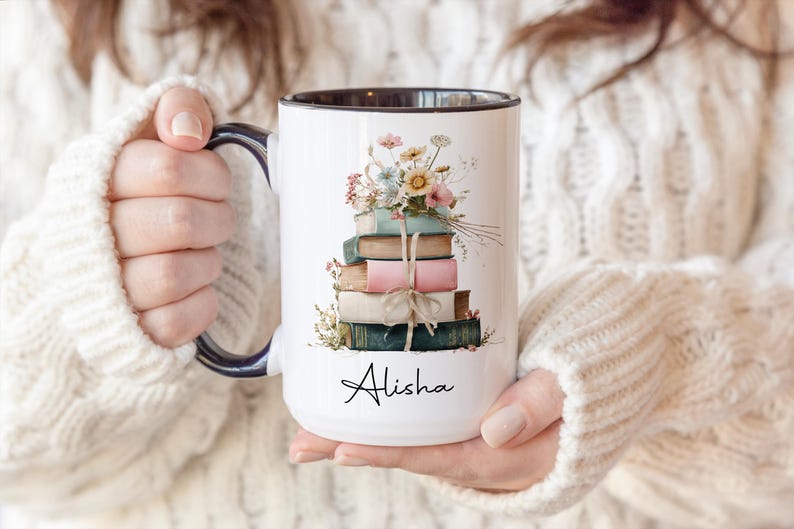 May include: White ceramic mug with a black rim and handle, featuring a stack of books tied with twine and topped with flowers. The name "Alisha" is written in script on the front. The mug is held by a person wearing a white sweater.