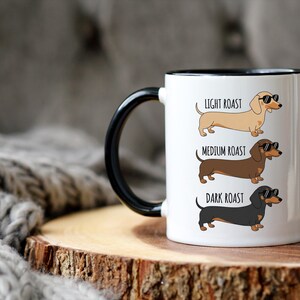 May include: White ceramic coffee mug with a black handle and rim. The mug features illustrations of dachshunds labeled "Light Roast," "Medium Roast," and "Dark Roast." Each dog wears sunglasses. The mug sits on a wooden surface.
