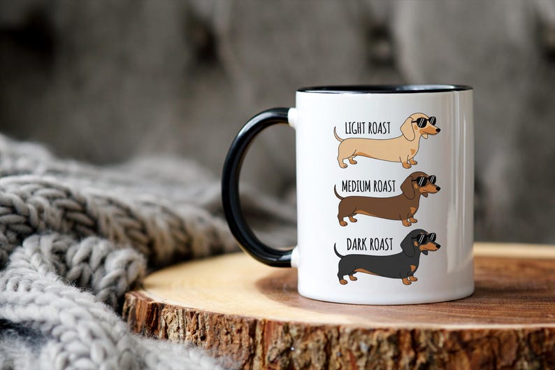 May include: White ceramic coffee mug with a black handle and rim. The mug features illustrations of dachshunds labeled "Light Roast," "Medium Roast," and "Dark Roast." Each dog wears sunglasses. The mug sits on a wooden surface.