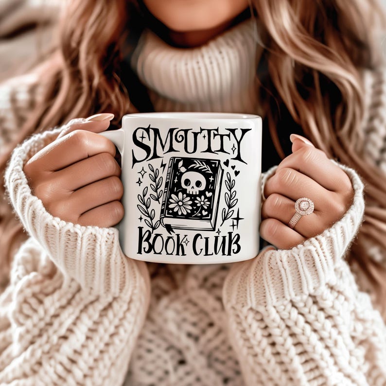 K&ouml;nnte beinhalten: Wei&szlig;e Keramiktasse mit dem schwarzen Schriftzug "SMUTTY BOOK CLUB". Die Tasse zeigt eine schwarze Illustration eines Buches mit einem Totenkopf, Blumen und botanischen Elementen. Die Tasse wird von einer Person gehalten, die einen cremefarbenen Pullover tr&auml;gt.
