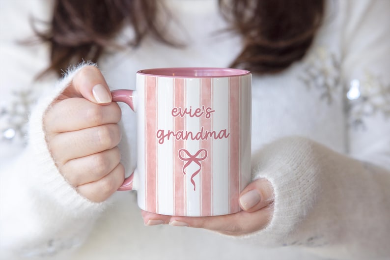 May include: A white and pink striped ceramic mug with the words "evie's grandma" and a bow graphic. The mug has a pink interior and handle. The mug is held by a person wearing a white sweater.