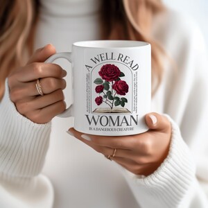 May include: White ceramic mug featuring a design with red roses and the text "A Well Read Woman Is A Dangerous Creature." The mug has a handle and is held by a person wearing a white sweater.