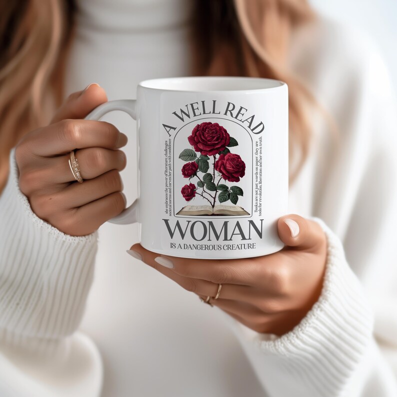 May include: White ceramic mug featuring a design with red roses and the text "A Well Read Woman Is A Dangerous Creature." The mug has a handle and is held by a person wearing a white sweater.