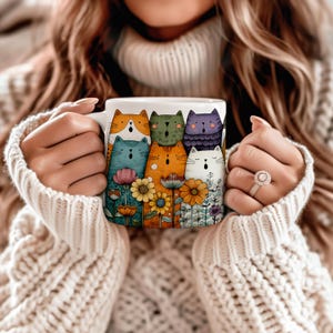Może przedstawiać: Biały ceramiczny kubek z kolorową ilustracją kresk&oacute;wkowych kot&oacute;w i kwiat&oacute;w. Koty są w r&oacute;żnych kolorach, w tym pomarańczowym, zielonym, niebieskim i fioletowym. Kwiaty są ż&oacute;łte, r&oacute;żowe i białe. Kubek jest trzymany przez osobę w kremowym swetrze.