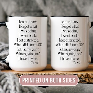 May include: Two white ceramic mugs with black handles, each printed with humorous text. The text reads "I came, I saw, I forgot what I was doing..." and is attributed to "Carol". The mugs are displayed on a wooden surface.
